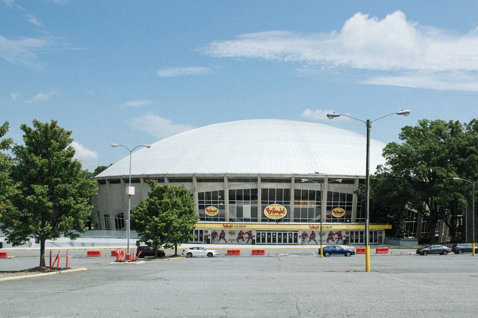 A large white stadium with a dome roof, surrounded by trees and parked cars.
