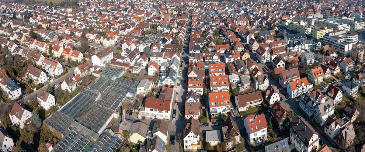 An aerial view of a densely populated residential area with numerous houses, a large greenhouse complex, and a railway track.