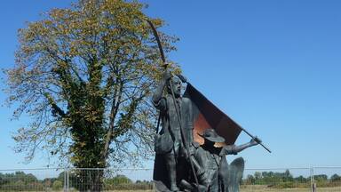 A bronze statue of a historical figure with a flag, set in a circular base with a tree and open field in the background.