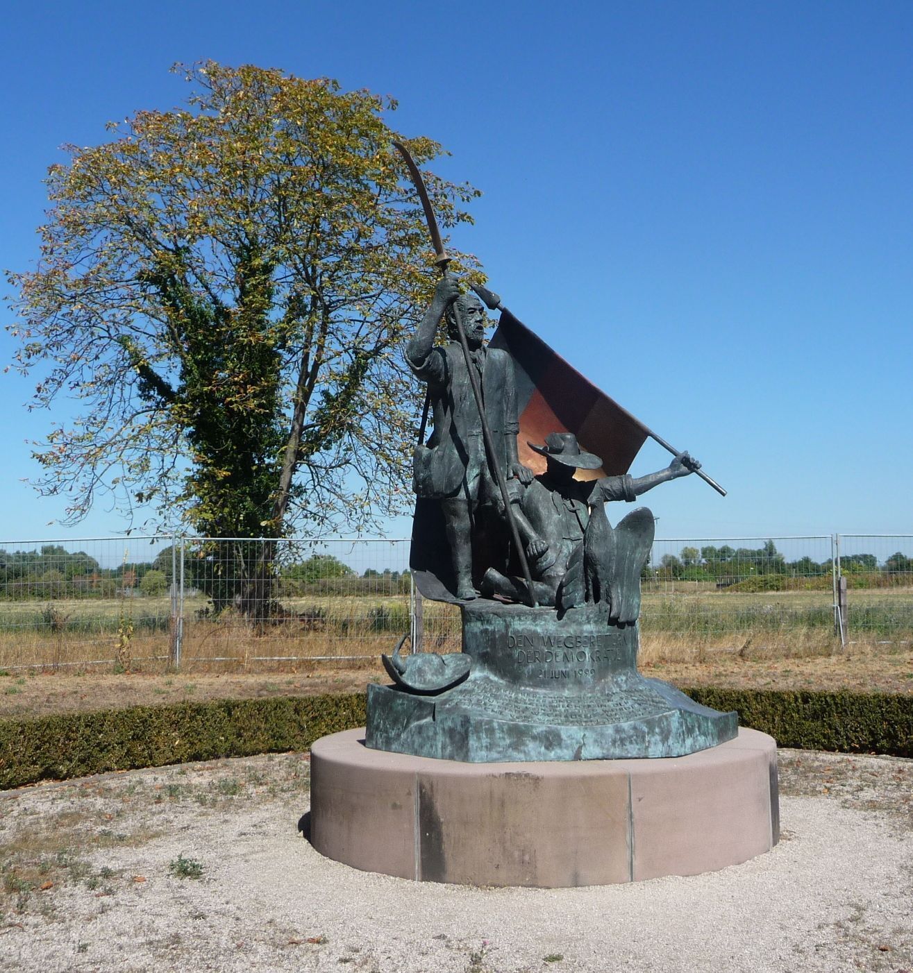 A bronze statue of a historical figure with a flag, set in a circular base with a tree and open field in the background.