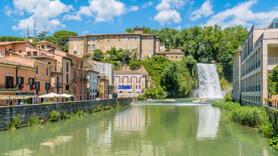A river with a waterfall, historic buildings, and lush greenery.