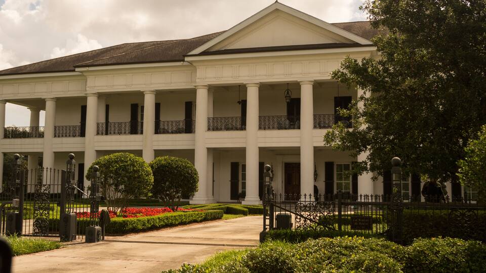 A large white building with columns and a portico, surrounded by well-maintained gardens and a fence.