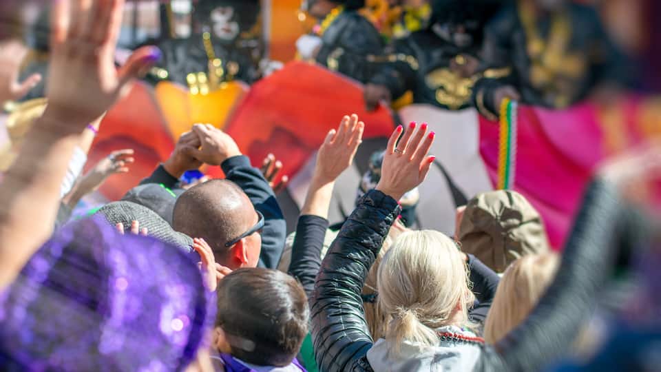 A crowd of people with raised hands, some wearing colorful clothing, and a person in the background with a red and gold outfit.
