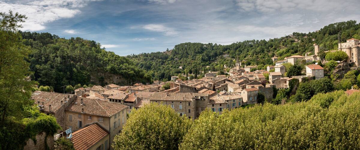 A historic town nestled in a valley with terracotta-roofed buildings, a prominent castle on a hill, and lush green hills in the background.