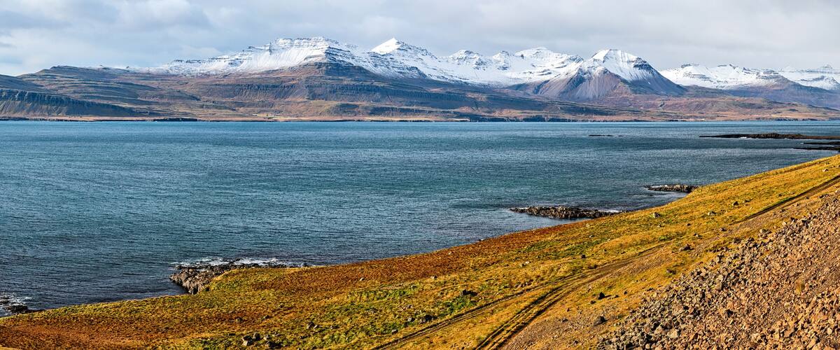 A coastal landscape with a rocky shoreline, a calm sea, and snow-capped mountains in the background.