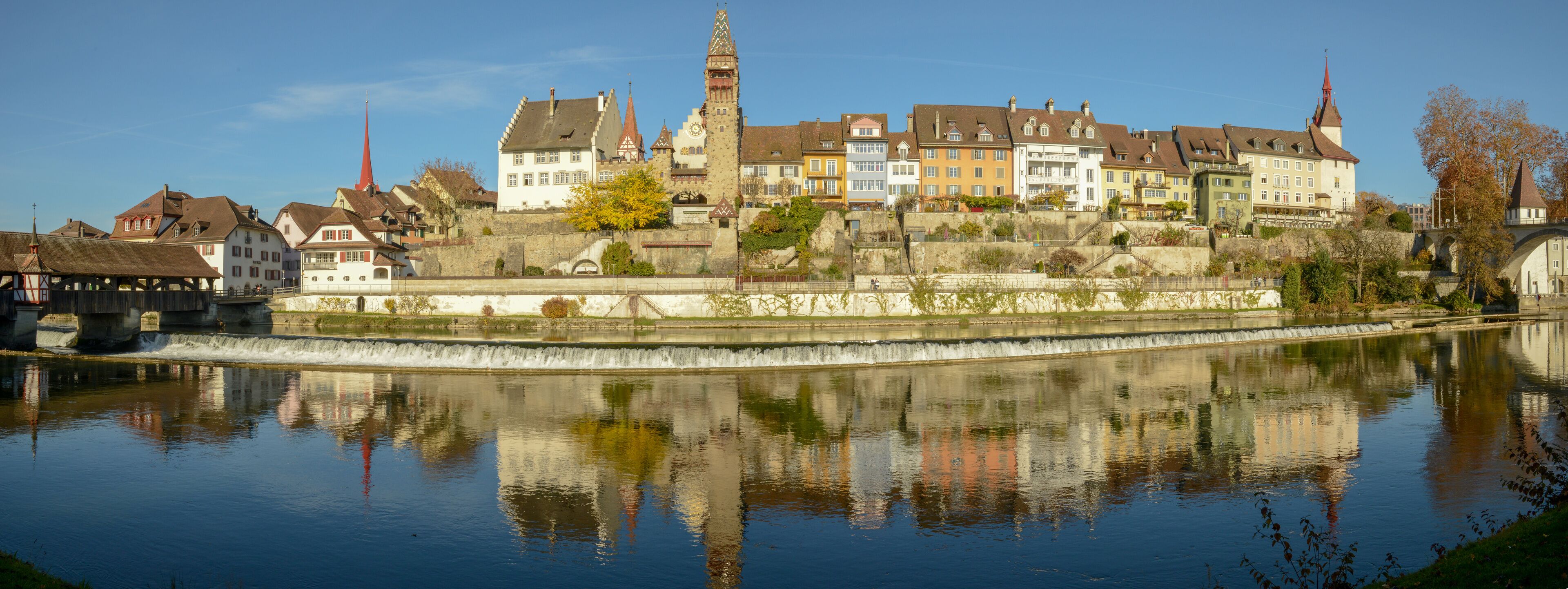 A historic town with a river, reflecting buildings and a church tower.