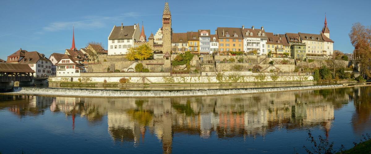 A historic town with a river, reflecting buildings and a church tower.