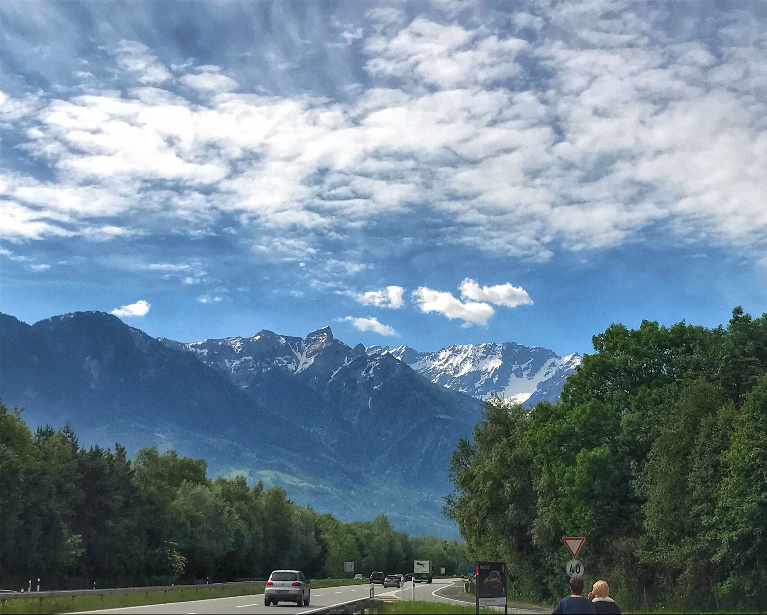 A road with vehicles, a speed limit sign, and a mountain range with snow-capped peaks under a partly cloudy sky.