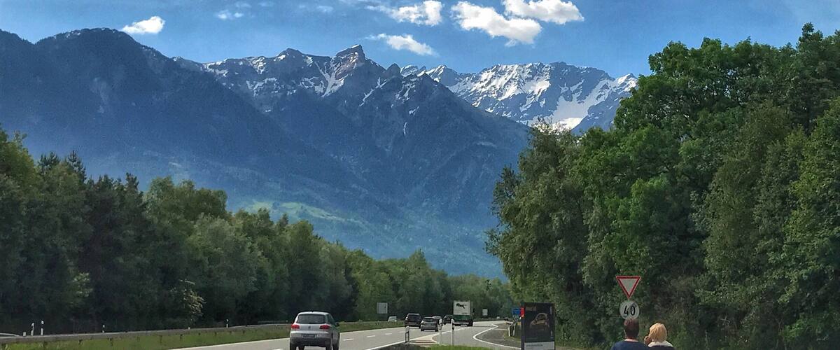 A road with vehicles, a speed limit sign, and a mountain range with snow-capped peaks under a partly cloudy sky.