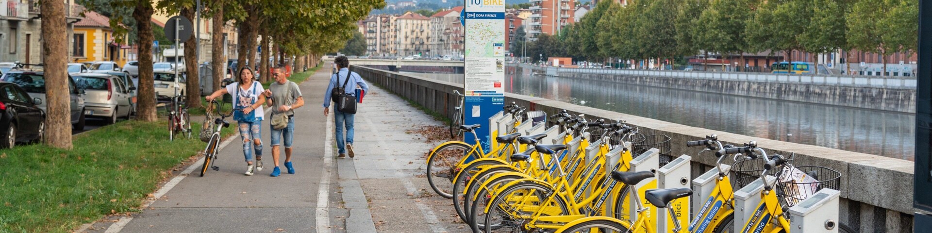 Uma estação de aluguel de bicicletas com bicicletas amarelas, um rio e edifícios ao fundo.