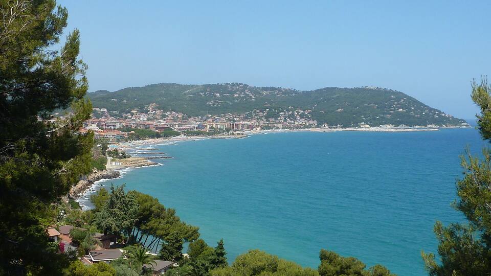 A coastal town with a clear blue sea, surrounded by hills and dense greenery.