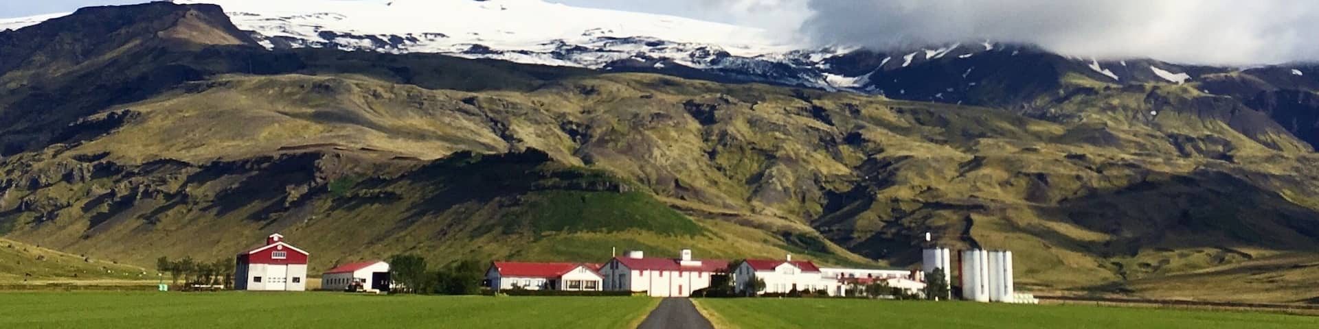 A rural landscape with a road leading to a cluster of white buildings with red roofs, snow-capped mountains in the background, and a partly cloudy sky.