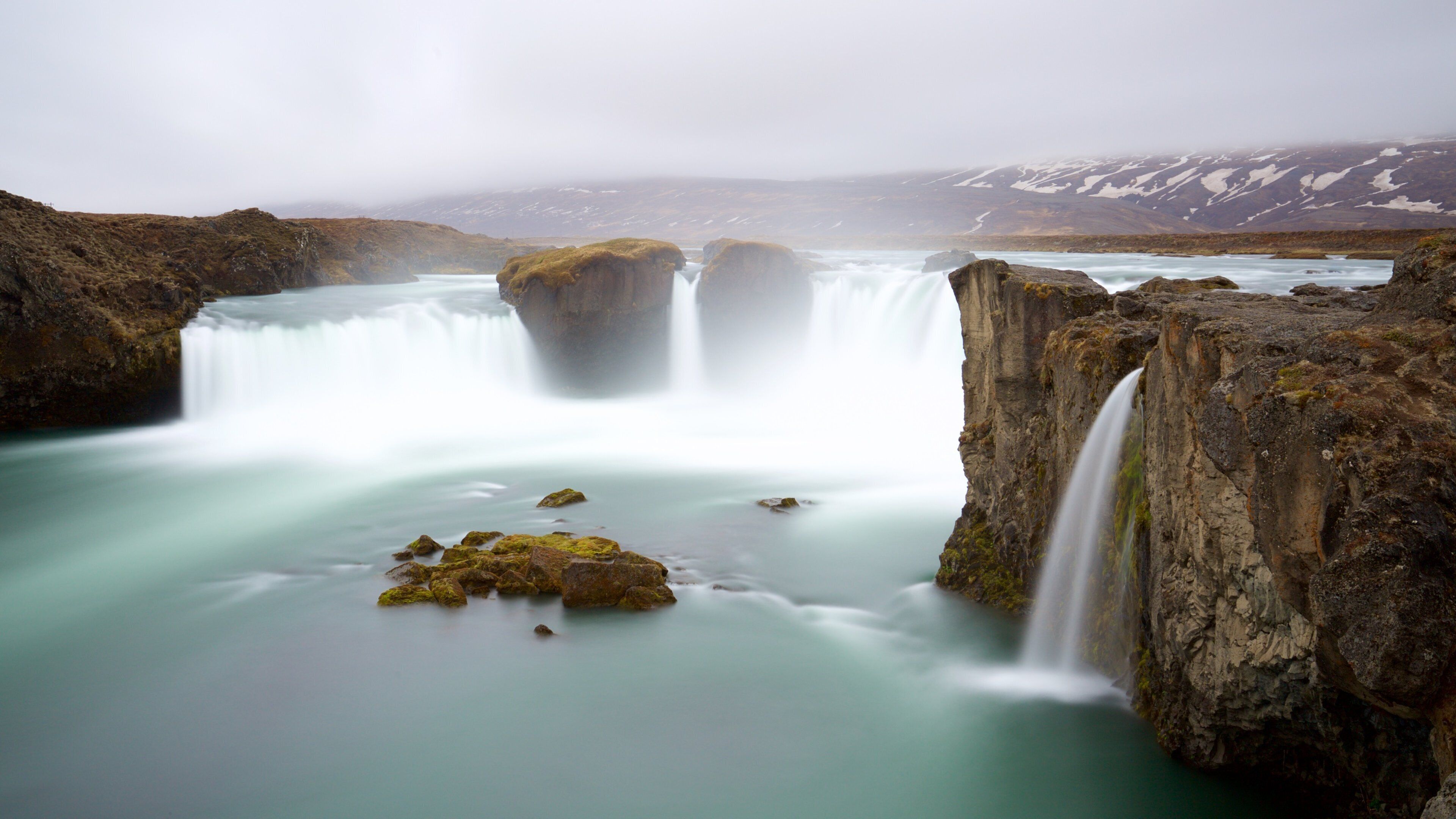 A waterfall cascading over rocky cliffs into a calm pool of water, surrounded by a mountainous landscape.