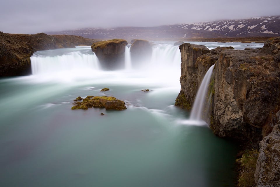 A waterfall cascading over rocky terrain with a misty atmosphere and distant snow-capped mountains.
