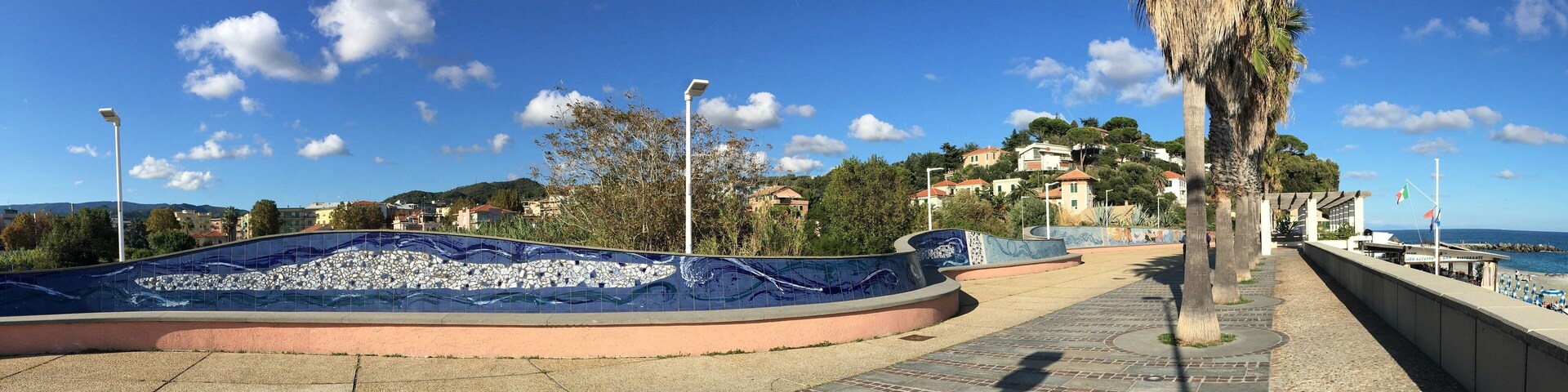 A coastal promenade with a mosaic pool, palm trees, and buildings on a hillside.