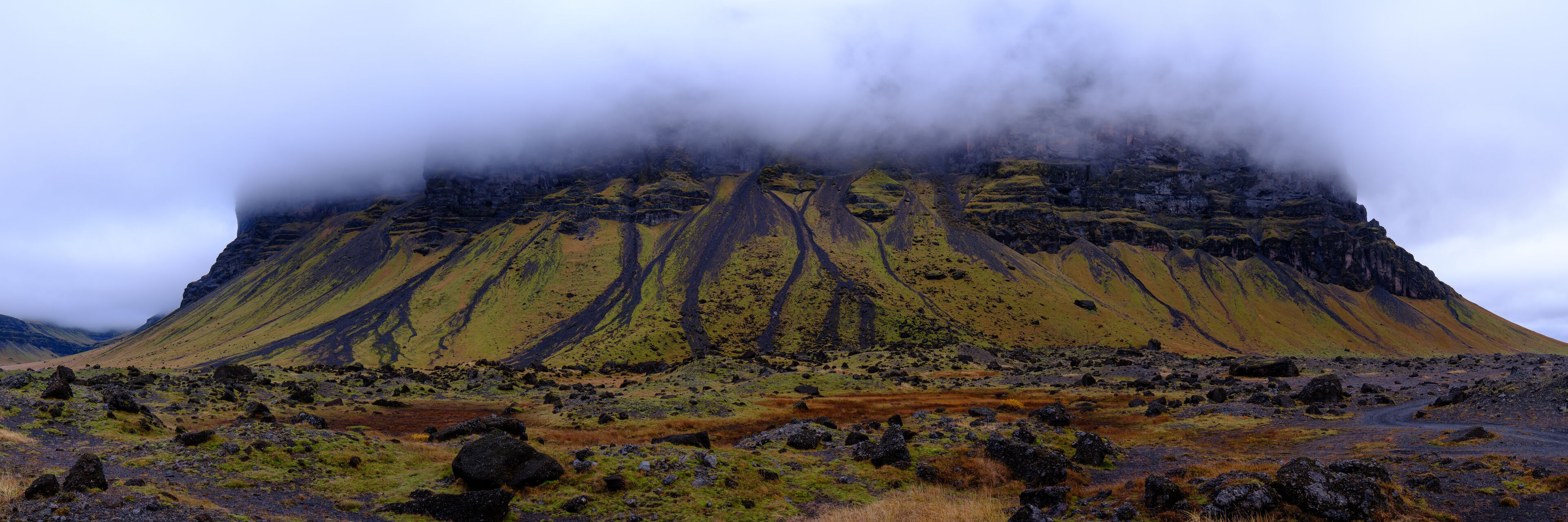 A rugged mountain landscape with a prominent volcanic cone partially obscured by clouds.
