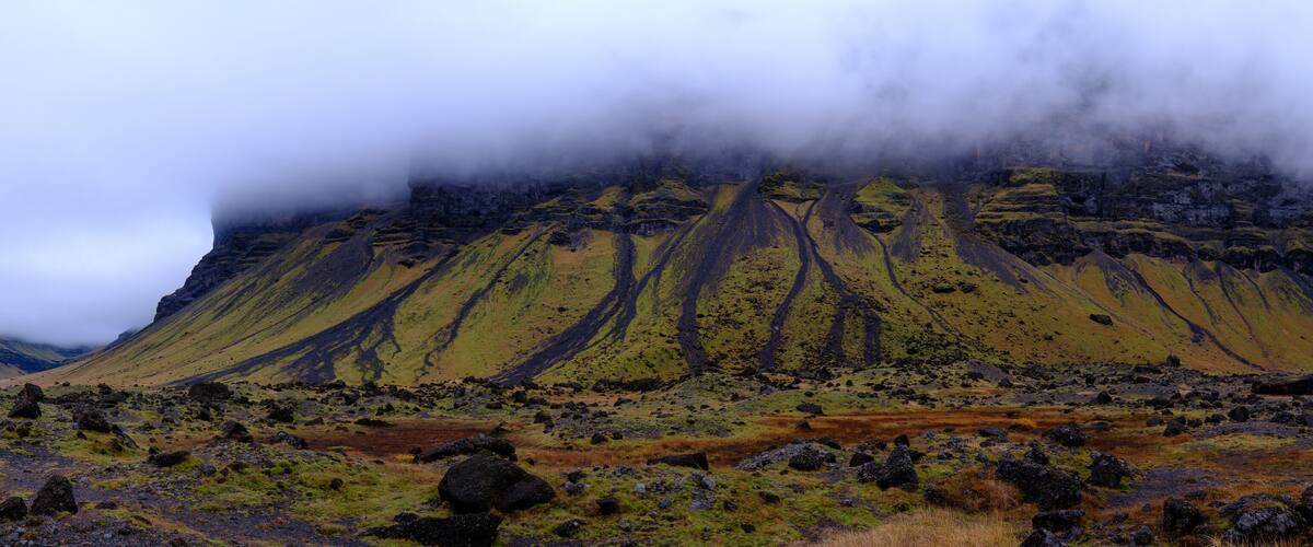 A rugged mountain landscape with a prominent volcanic cone partially obscured by clouds.