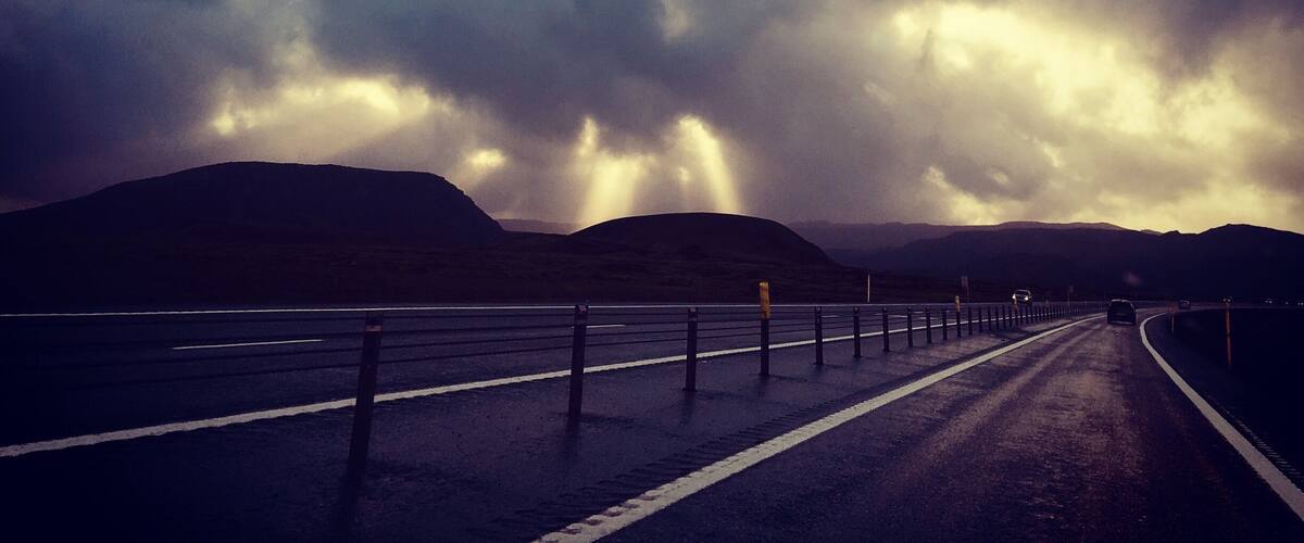 A road with a guardrail, a mountain range, and a partly cloudy sky with sunlight breaking through.