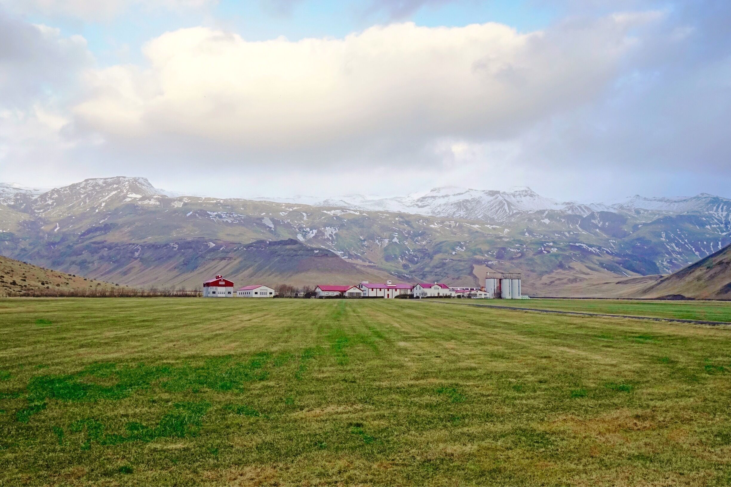 A rural landscape with a row of buildings and a field in front of snow-capped mountains.
