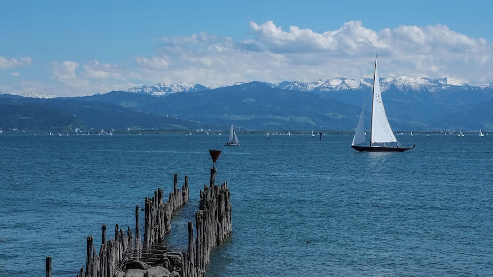 Eine felsige Küste mit einem hölzernen Steg, der sich in ruhiges Wasser erstreckt. In der Ferne sind Segelboote vor einer schneebedeckten Bergkulisse unter blauem Himmel zu sehen.