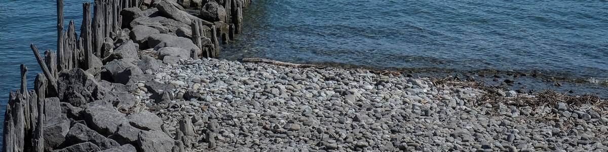 Eine felsige Küste mit einem hölzernen Steg, der sich in ruhiges Wasser erstreckt. In der Ferne sind Segelboote vor einer schneebedeckten Bergkulisse unter blauem Himmel zu sehen.