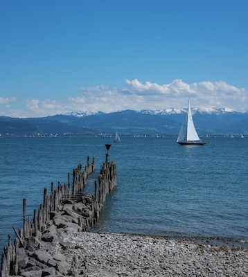 Eine felsige Küste mit einem hölzernen Steg, der sich in ruhiges Wasser erstreckt. In der Ferne sind Segelboote vor einer schneebedeckten Bergkulisse unter blauem Himmel zu sehen.