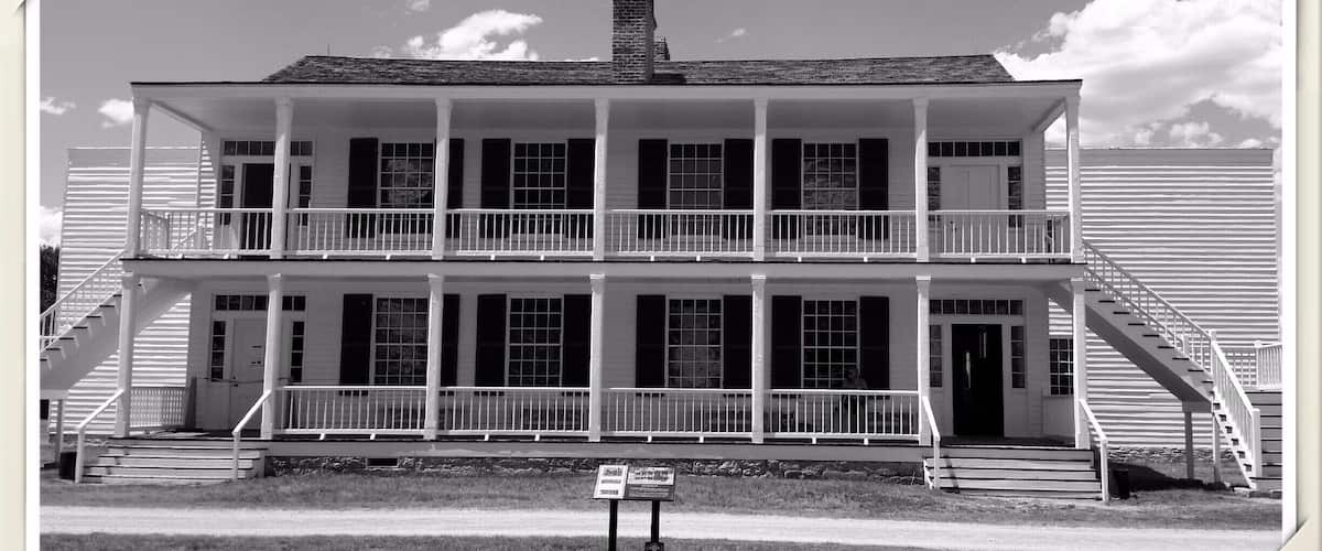 A two-story white colonial-style building with a central chimney, multiple windows, and a front porch with a staircase leading to the second floor.