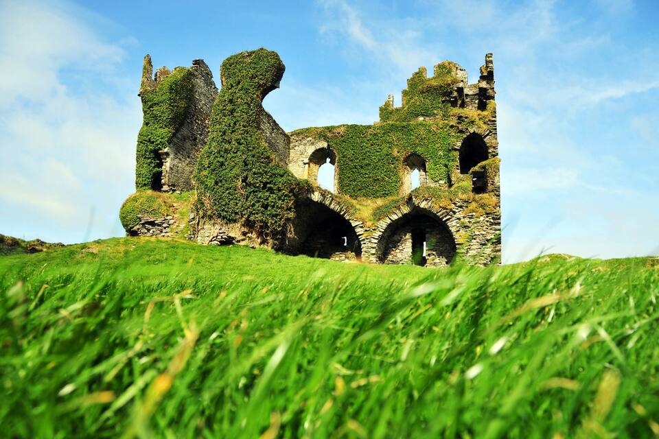 A historic castle ruins with ivy-covered walls and arches, set against a clear sky.