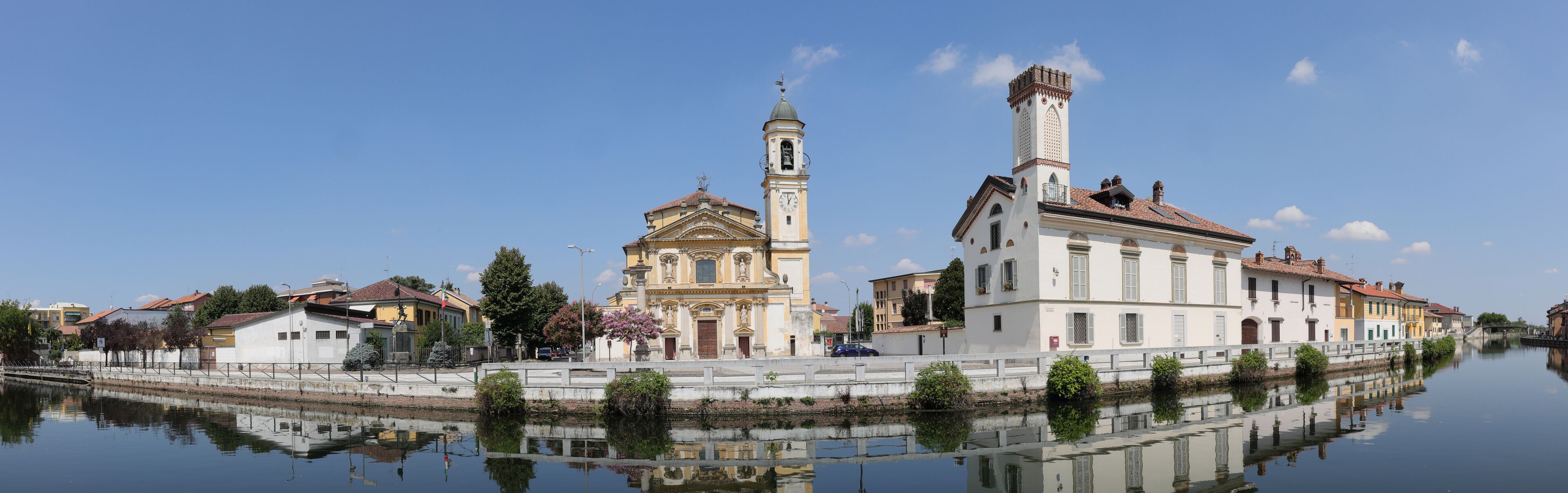 A historic town with a prominent church, a bell tower, and a canal reflecting the architecture.