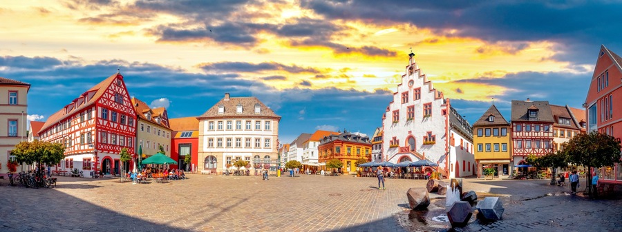 A cobblestone square with traditional European architecture, a fountain, and a sunset sky.