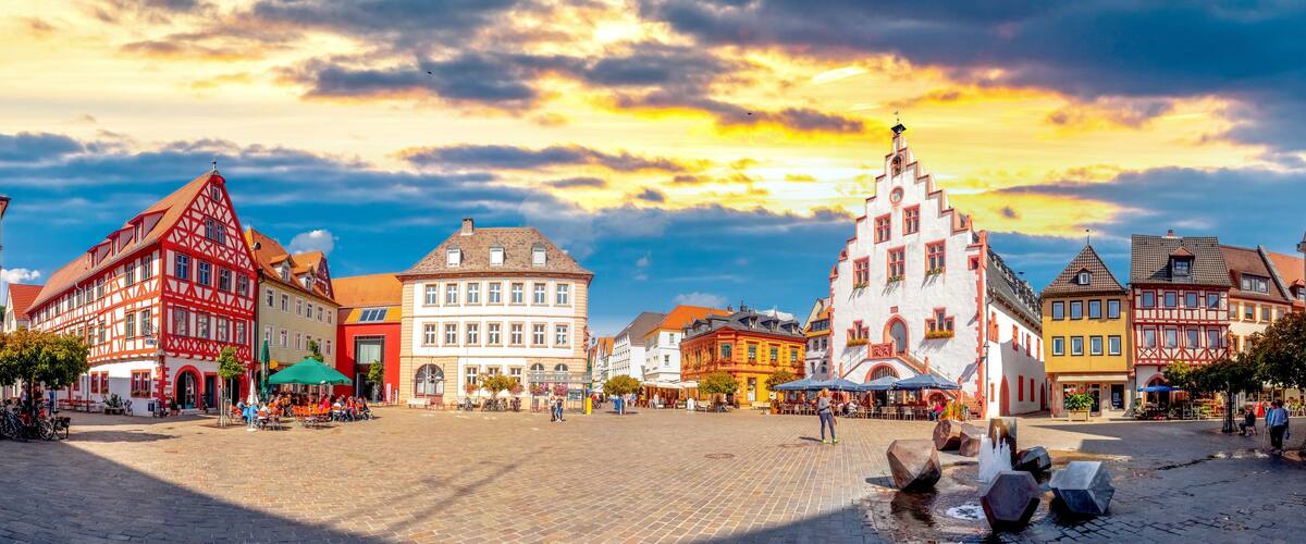 A cobblestone square with traditional European architecture, a fountain, and a sunset sky.