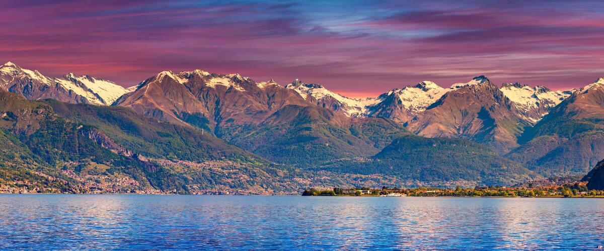 A mountain range with snow-capped peaks, a lake in the foreground, and a town nestled at the base of the mountains.