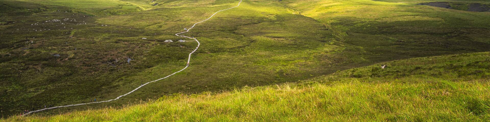 Winding wooden path of Cuilcagh Mountain Park boardwalk, illuminated by patches of sunlight in the valley below with dramatic sky, Northern Ireland
