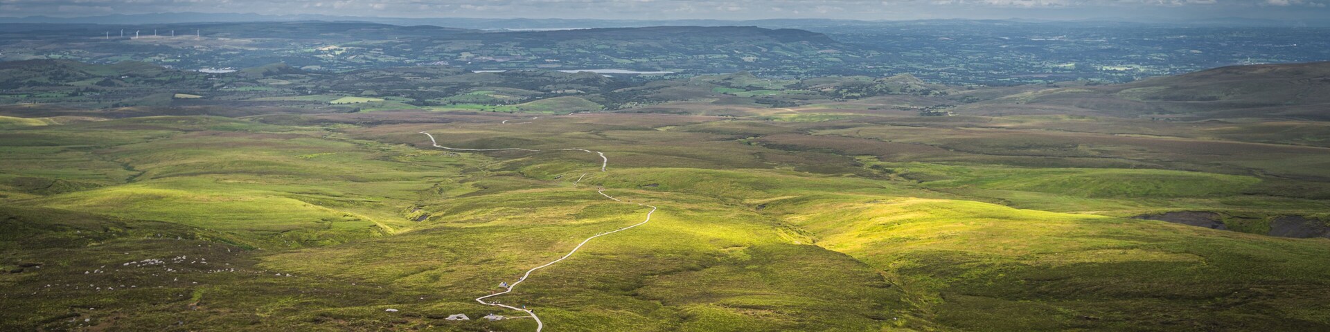 Winding wooden path of Cuilcagh Mountain Park boardwalk, illuminated by patches of sunlight in the valley below with dramatic sky, Northern Ireland