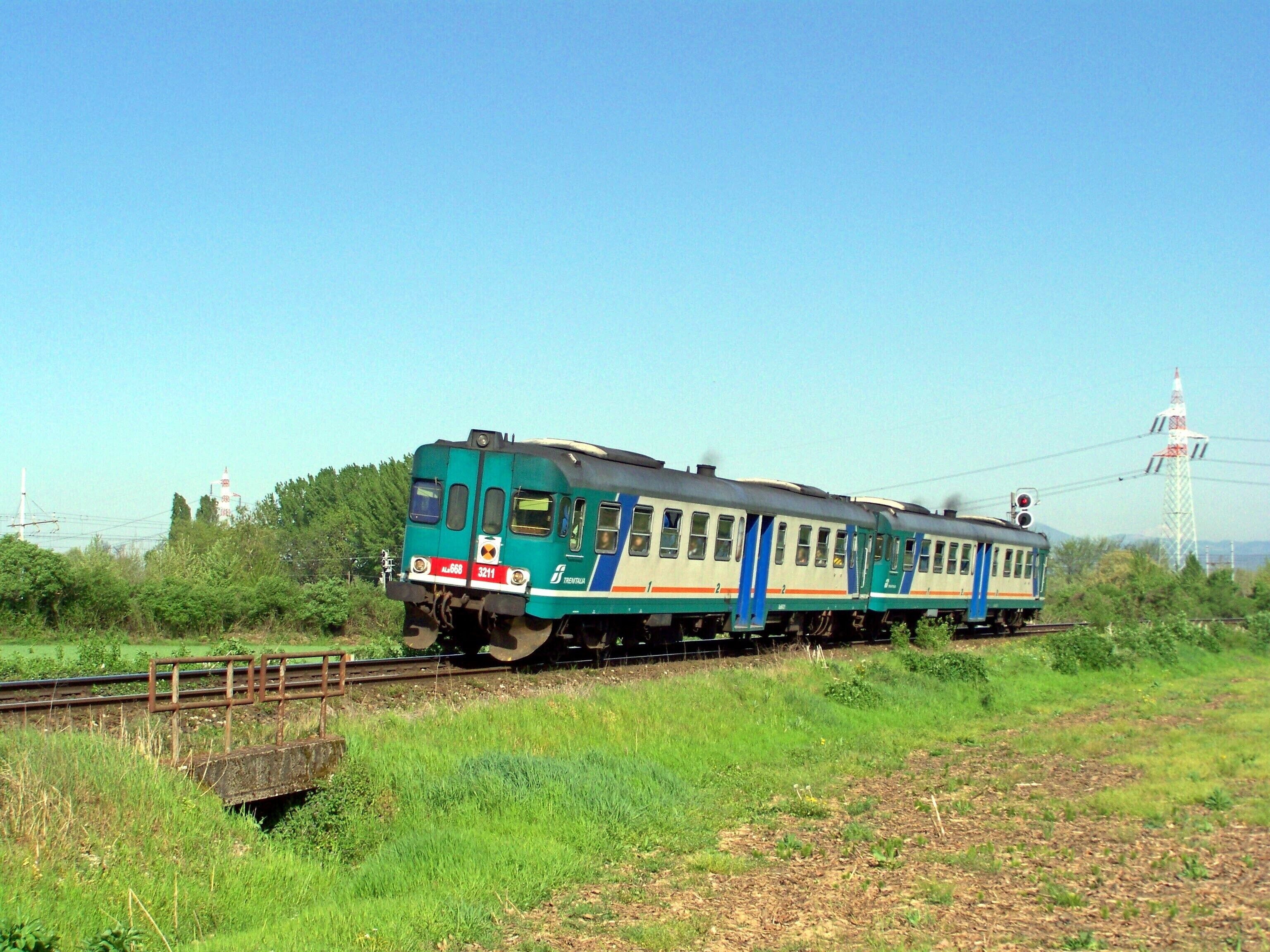 A green and white train on tracks with a bridge and greenery in the background.
