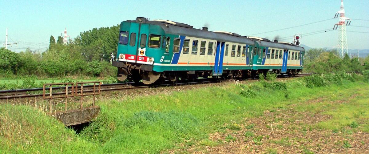 A green and white train on tracks with a bridge and greenery in the background.