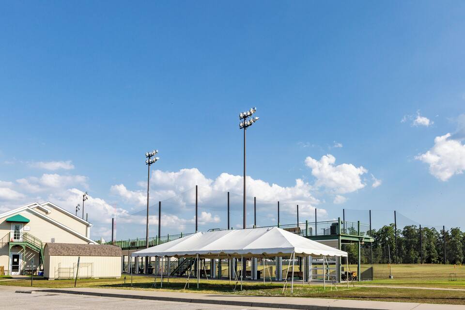 A sports field with a white tent, floodlights, and a building with a green roof.