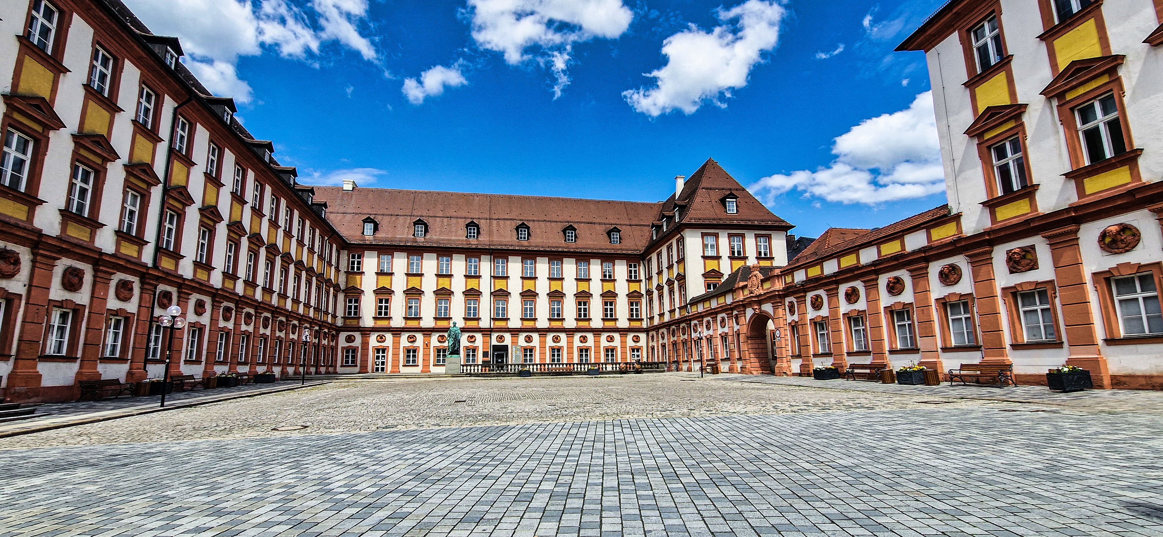 A historic courtyard with a central building featuring a red-tiled roof and yellow accents, surrounded by classical architecture.