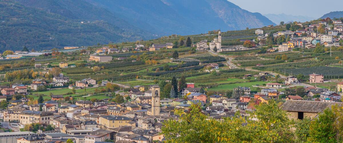 A town nestled in a valley with vineyards, a church, and mountains in the background.