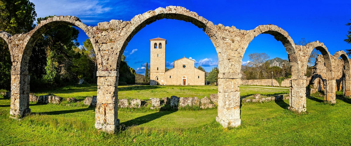 Ancient stone arches, a church with a bell tower, and a grassy field.