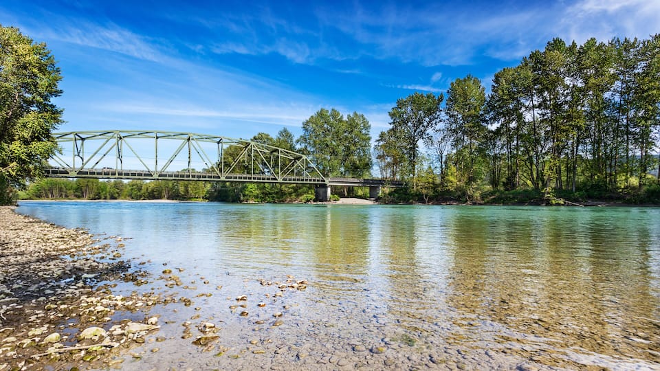 A green metal bridge over a river with clear water, surrounded by trees and pebbles on the shore.