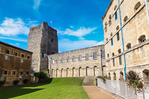 A historic stone building with a tower, a courtyard with a lawn, and a white fence with flowers.