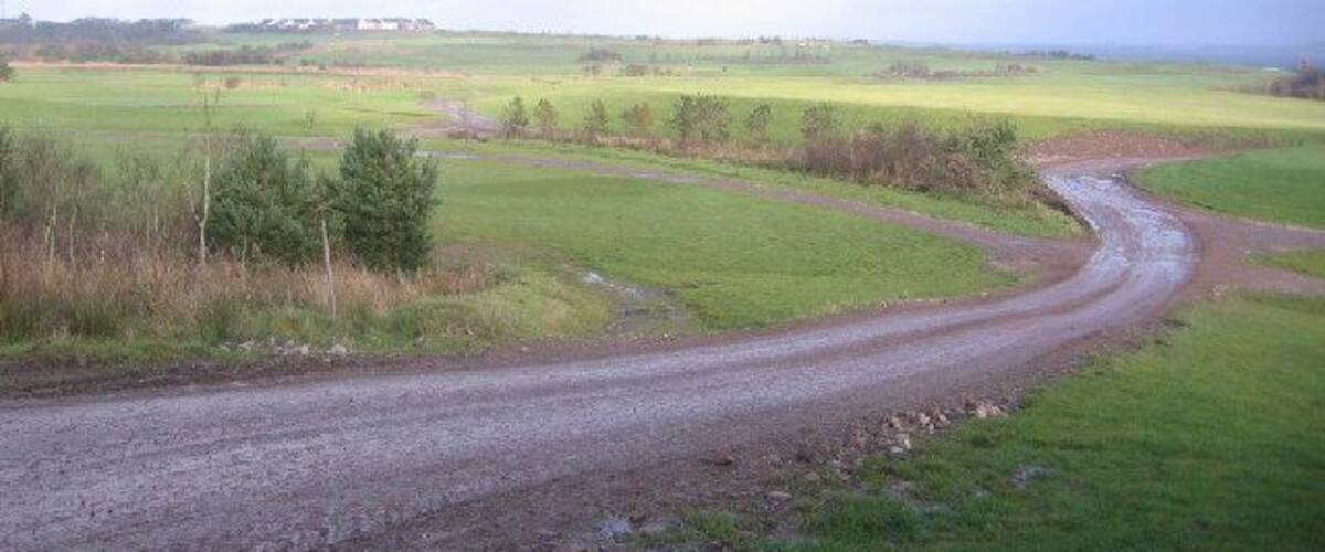 A rural dirt road curving through green fields with a distant building.