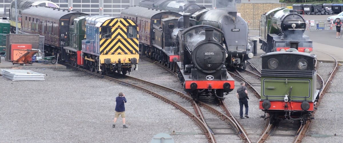 A steam locomotive at a railway station with multiple tracks and a modern building in the background.