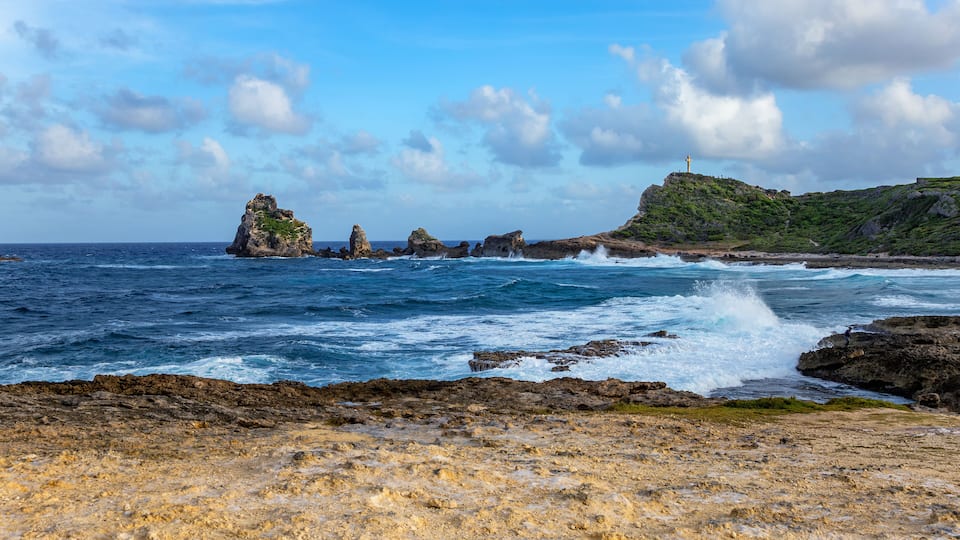 A rocky coastline with waves crashing against the shore, a lighthouse on a hill, and a clear blue sky.