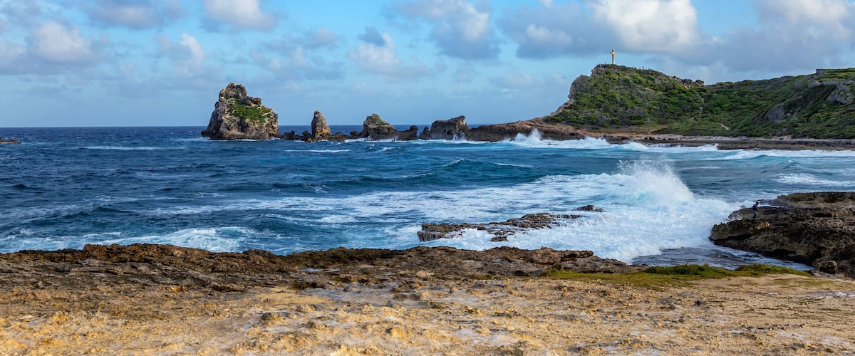 A rocky coastline with waves crashing against the shore, a lighthouse on a hill, and a clear blue sky.