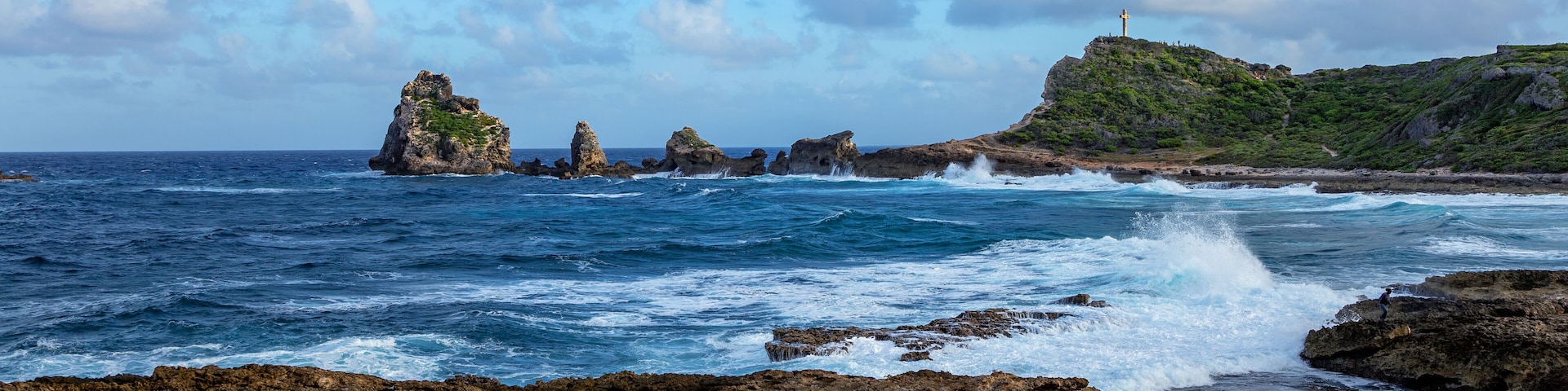 A rocky coastline with waves crashing against the shore, a lighthouse on a hill, and a clear blue sky.