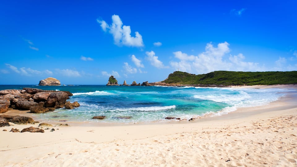 A beach with clear turquoise waters, rocky outcrops, and a distant lighthouse.