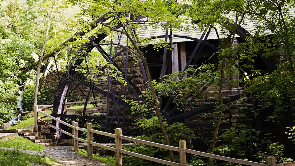 A stone water wheel surrounded by lush greenery and a wooden fence.