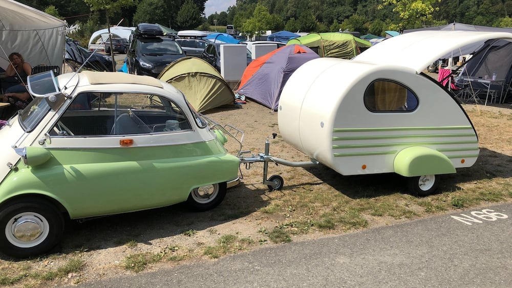 Ein alter Wohnmobilwagen steht neben einem grünen Auto, umgeben von Zelten und anderen Fahrzeugen auf einem Campingplatz.